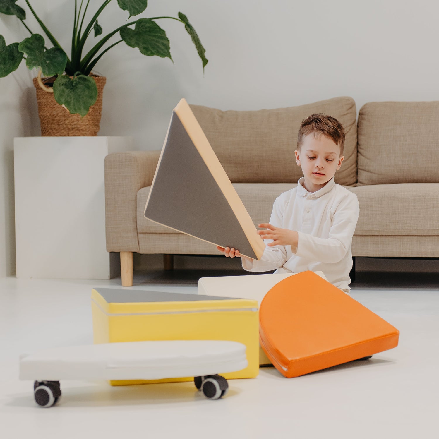 Child playing with geometric shapes on a white floor with a beige couch and plant in the background